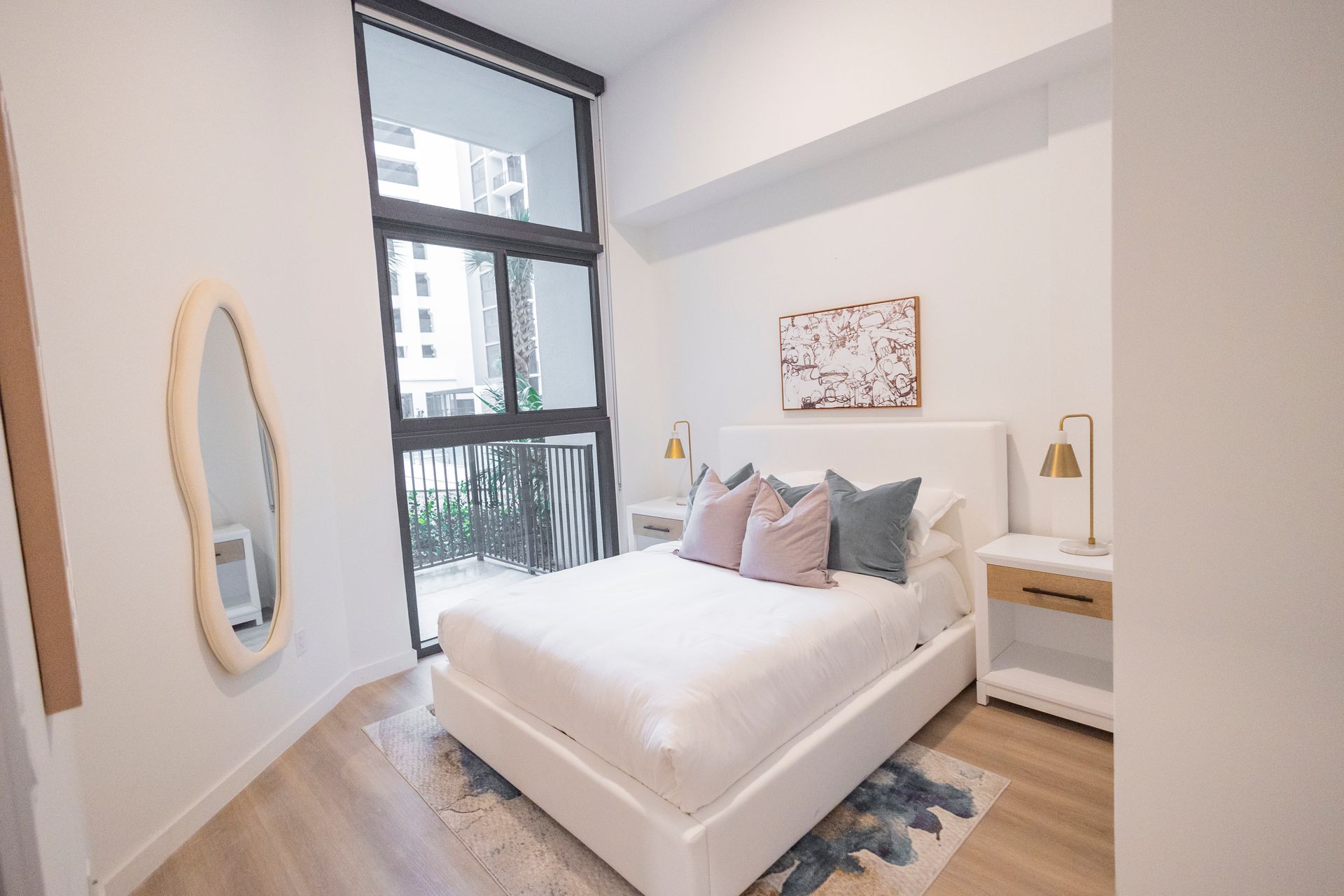 Bright modern bedroom with white bed, gray pillows, wood floor, and large window with black blinds at The Arcadian in Fort Lauderdale