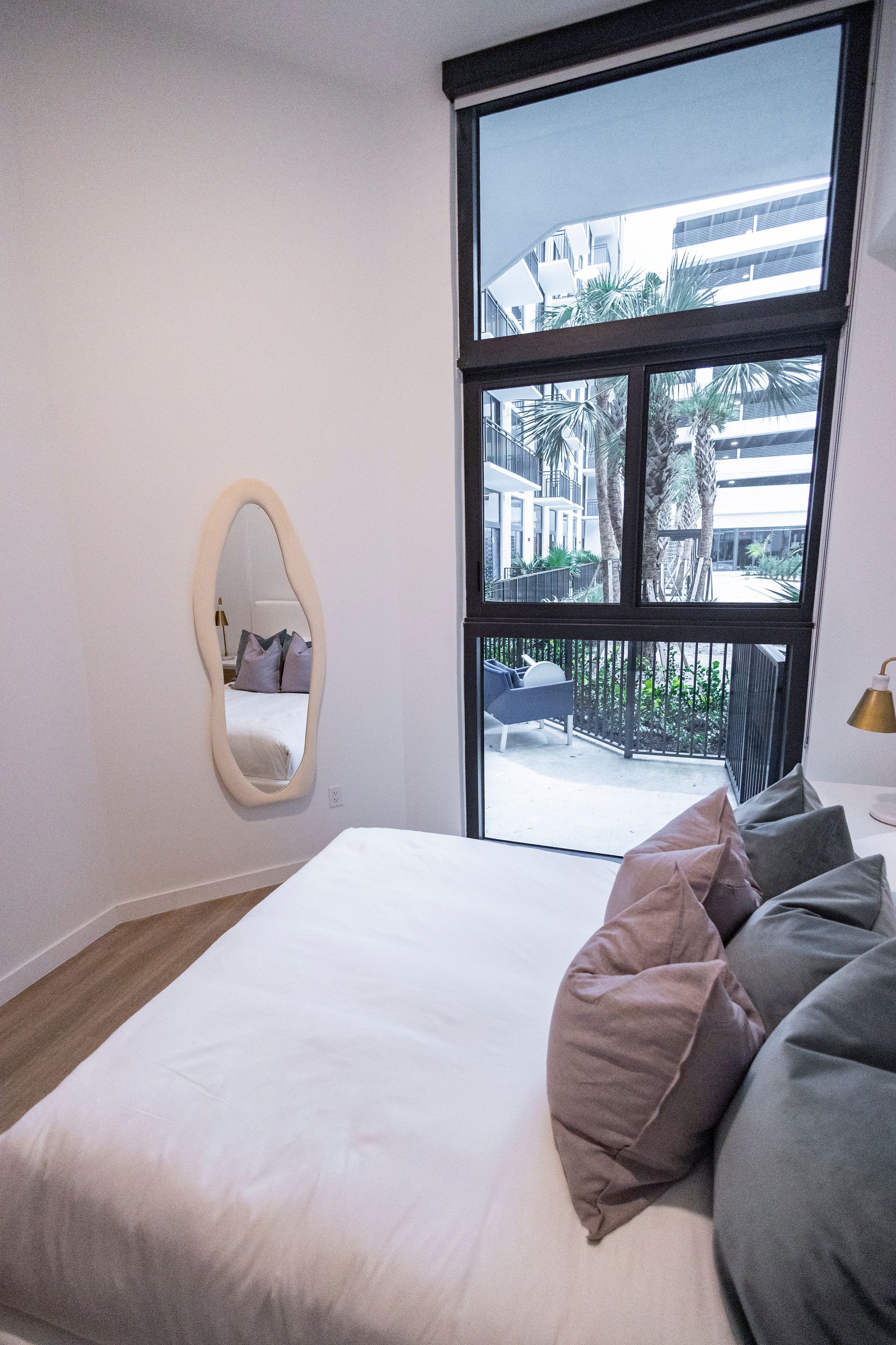 Bright bedroom with a bed, oval wall mirror, and large window overlooking a balcony at The Arcadian in Fort Lauderdale