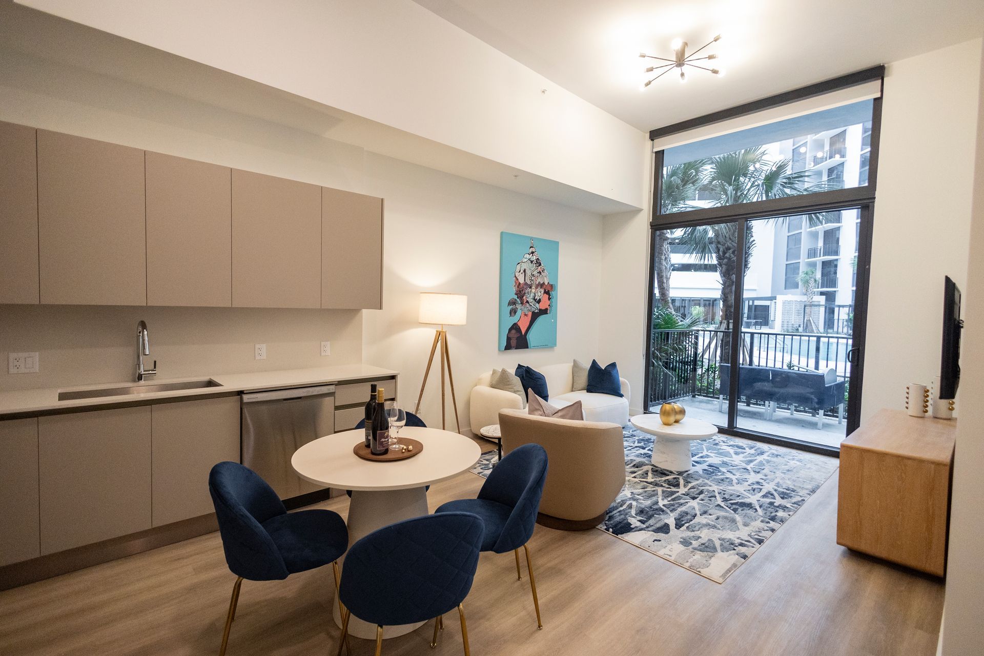 Modern apartment living room with blue chairs, round table, sofa, wall art, and large window to a balcony at The Arcadian in Fort Lauderdale