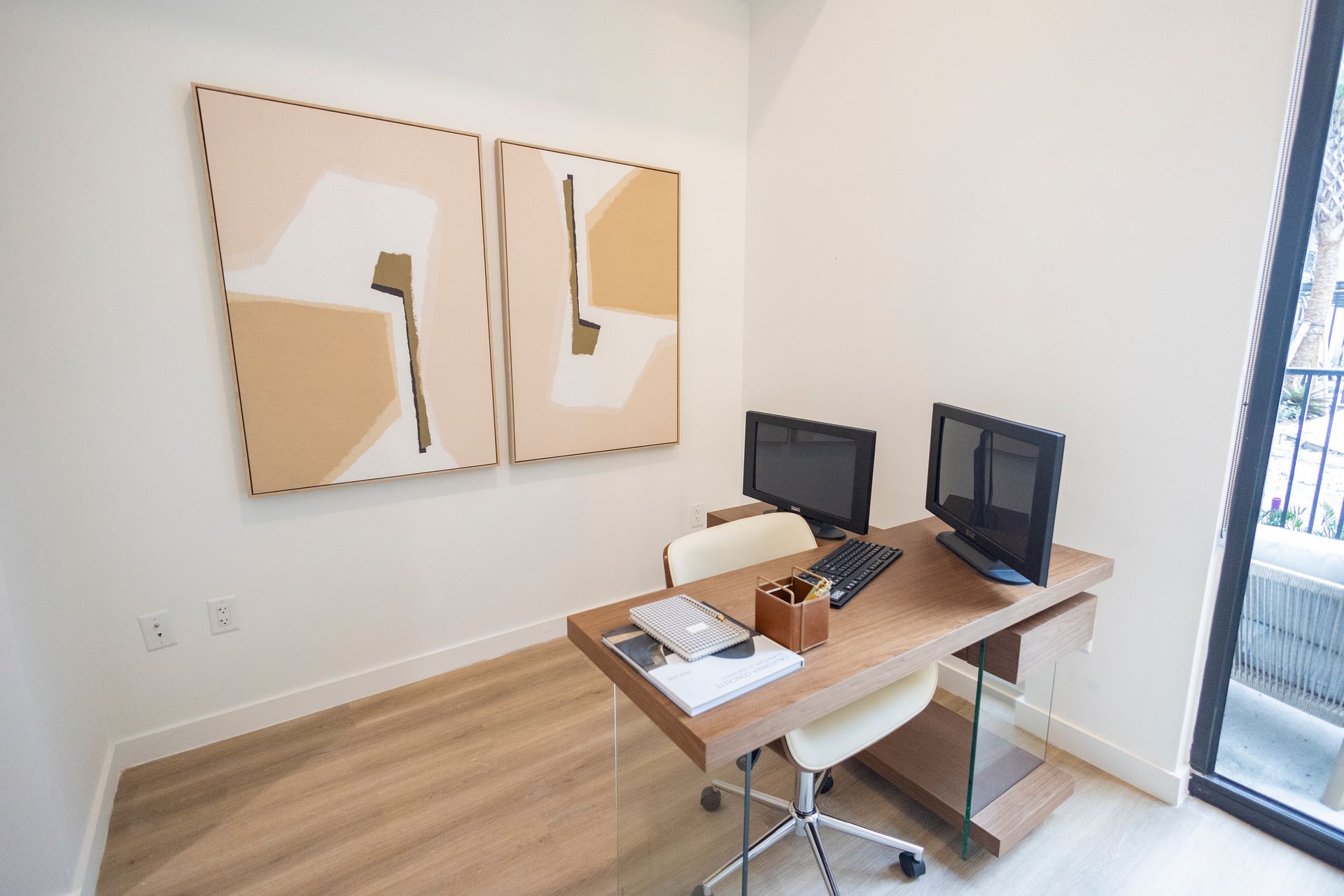 Modern home office with a wooden desk, dual monitors, beige chair, and abstract wall art at The Arcadian in Fort Lauderdale