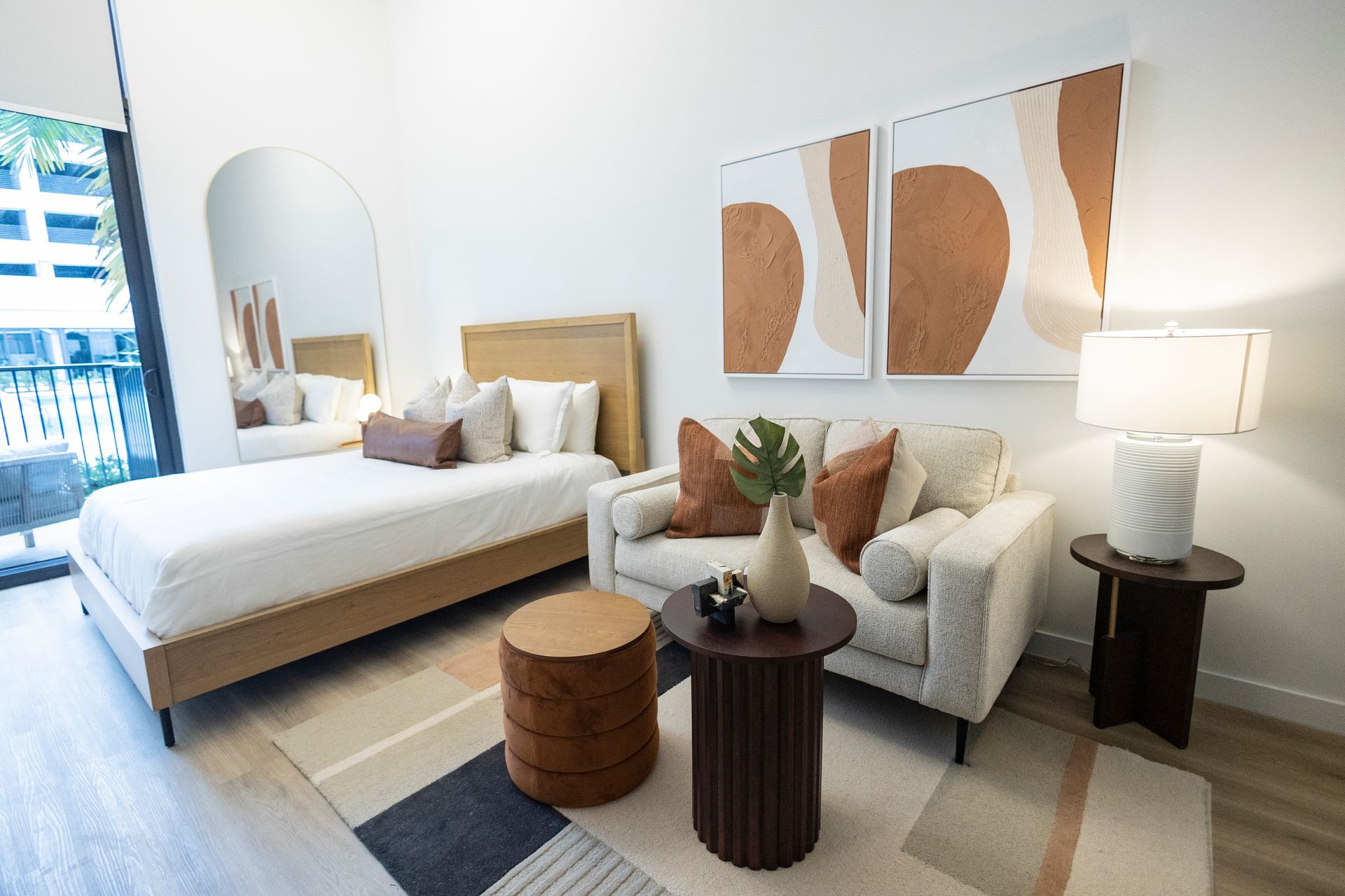 Bright modern bedroom with a bed, armchair, wooden tables, lamp, wall art, and patterned rug at The Arcadian