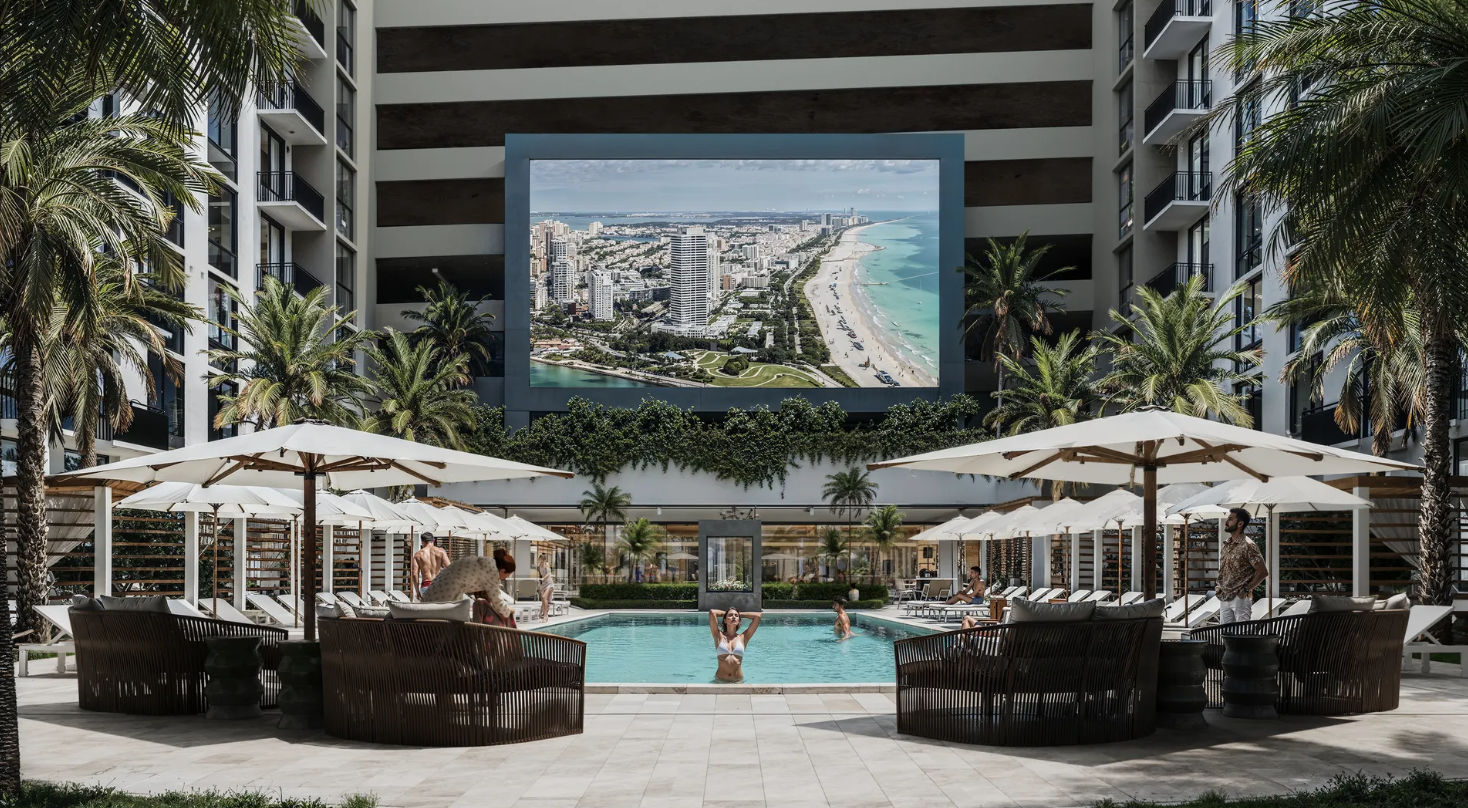Resort courtyard with a pool, white umbrellas, palm trees, and a large ocean-view screen backdrop at The Arcadian