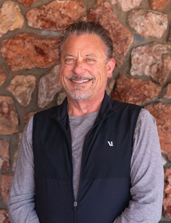 Smiling man in a dark vest standing against a stone wall