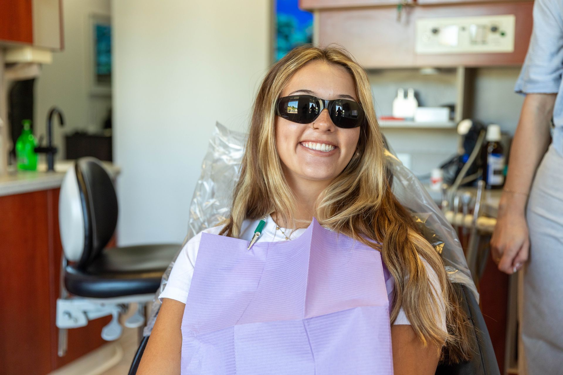 Smiling person in sunglasses and a purple dental bib seated in a dentist chair