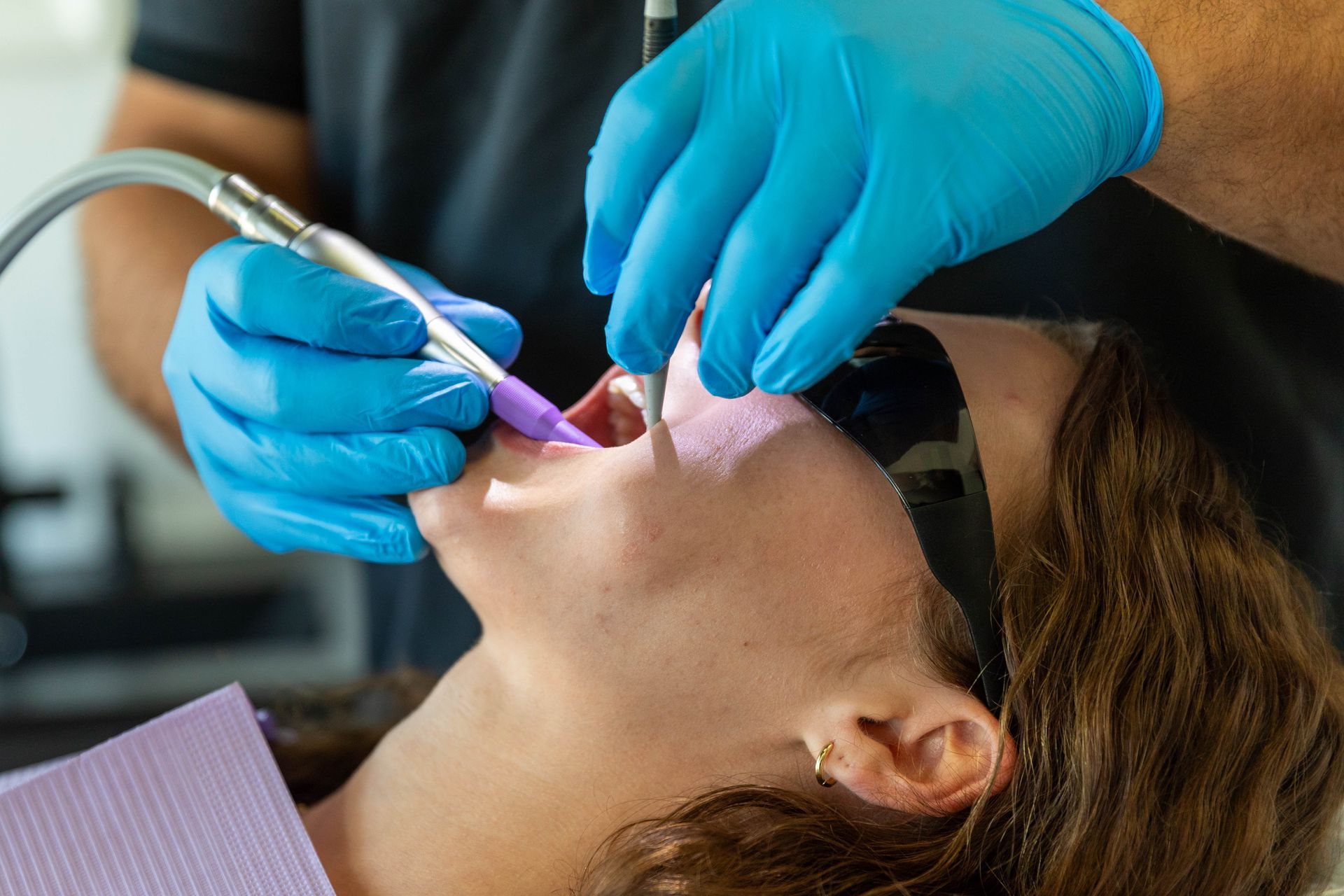 Dentist using tools on a patient’s teeth during a dental procedure, with blue gloves and a purple bib