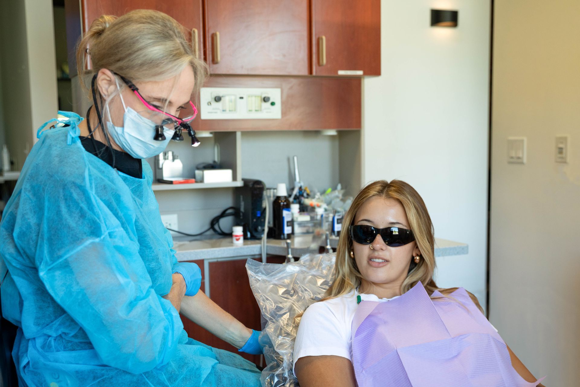 Dentist treating a patient in a dental chair, with both wearing protective gear and a purple bib.