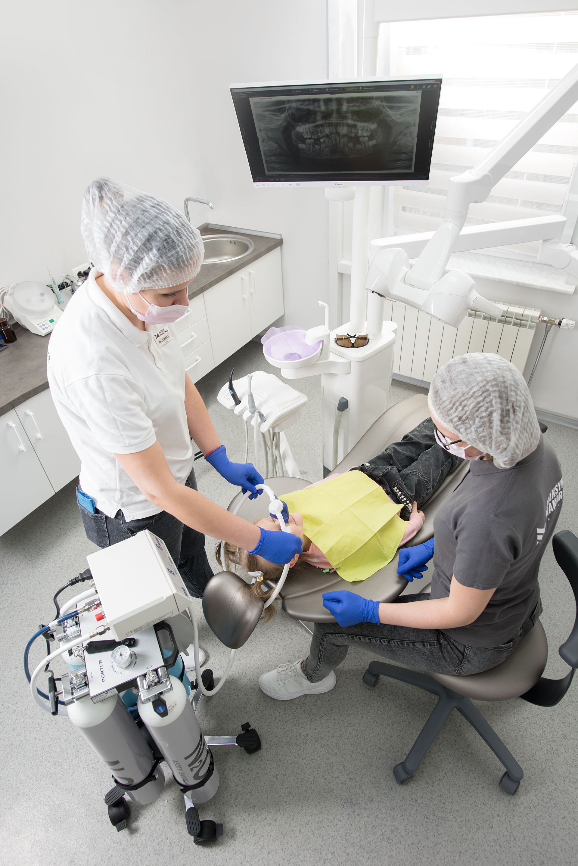 Dentist and assistant examining a patient in a bright clinic room, with a monitor and equipment nearby
