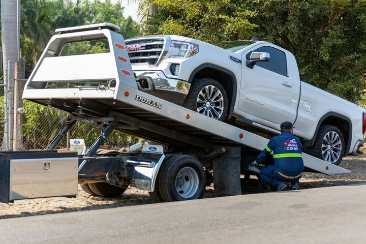 A tow truck operator in a blue uniform secures a white GMC pickup truck onto the flatbed of a tow truck outdoors.