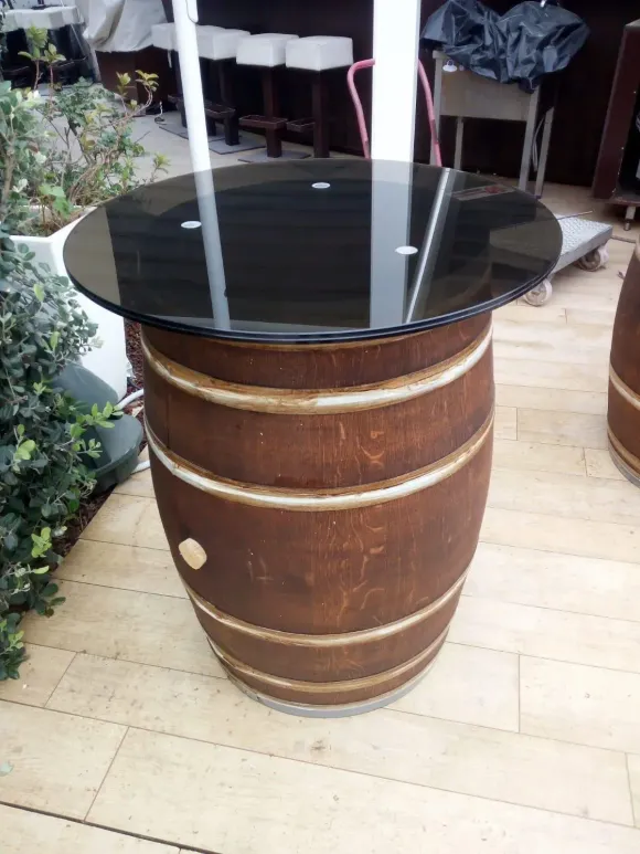 Tavolo rotondo con piano in vetro nero su base in legno a forma di botte, esposto all'aperto su un patio piastrellato.