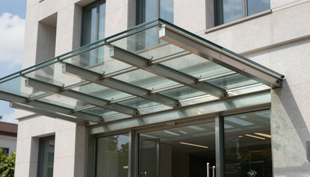 Tettoia in vetro sopra l'ingresso di un edificio con supporti metallici e facciata bianca.