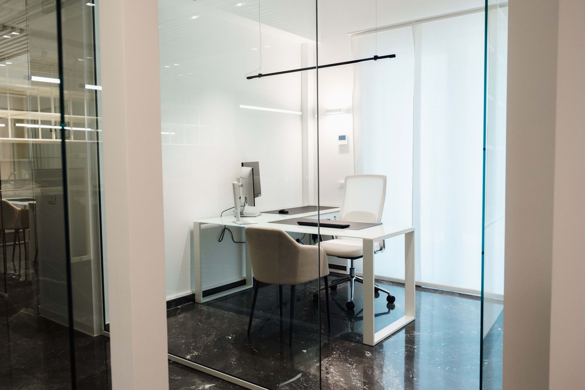 Ufficio con pareti in vetro, scrivania bianca, sedia e computer in un ambiente di lavoro moderno.
