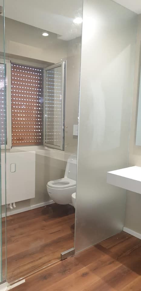Bagno moderno con doccia in vetro, WC bianco e pavimento in legno, illuminato da una finestra con tende.