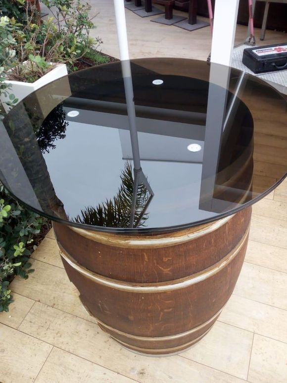 Tavolo con piano in vetro su base di legno ricavata da una botte, all'aperto, che riflette alberi e cielo.