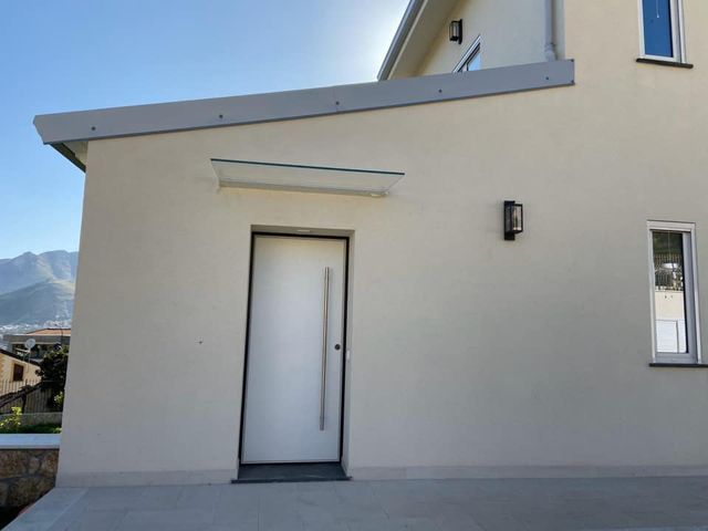 Esterno minimalista di una casa beige con porta d'ingresso bianca e piccola applique sotto un tetto spiovente.