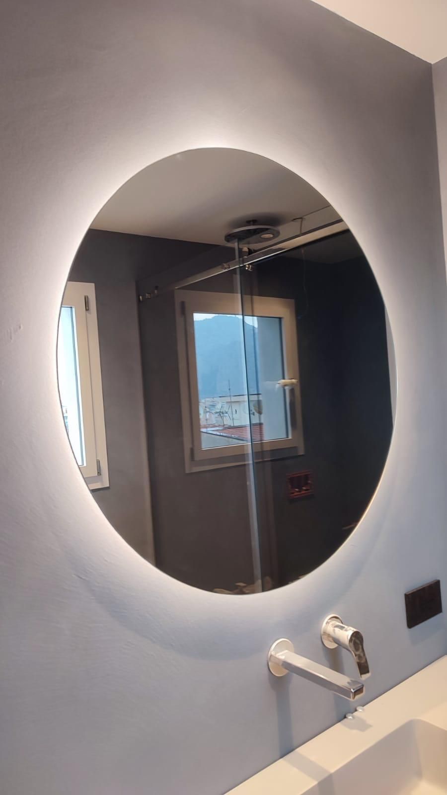 Specchio da bagno con retroilluminazione circolare sopra il lavabo e il rubinetto, che riflette la doccia e la finestra.