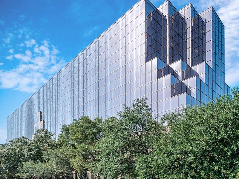 Modern glass office building with angular facade behind green trees under a blue sky
