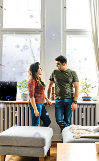 Two people talking by a window in a bright living room, with a white ottoman and sofa in the foreground.