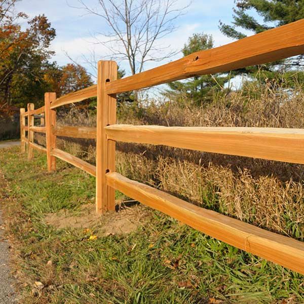 Wooden split-rail fence along a grassy field in autumn, with trees and dry brush in the background.