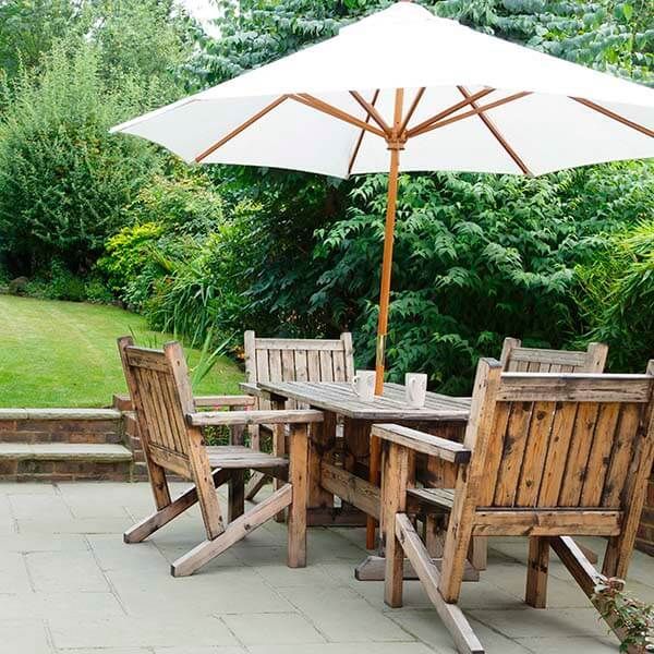 Wooden patio dining set with white umbrella on a stone terrace beside lush green shrubs and lawn.