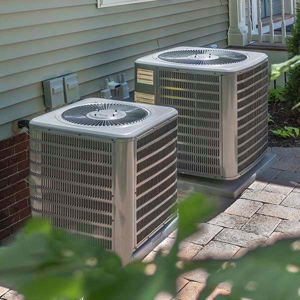 Two outdoor air conditioning units beside a house with siding and a brick wall