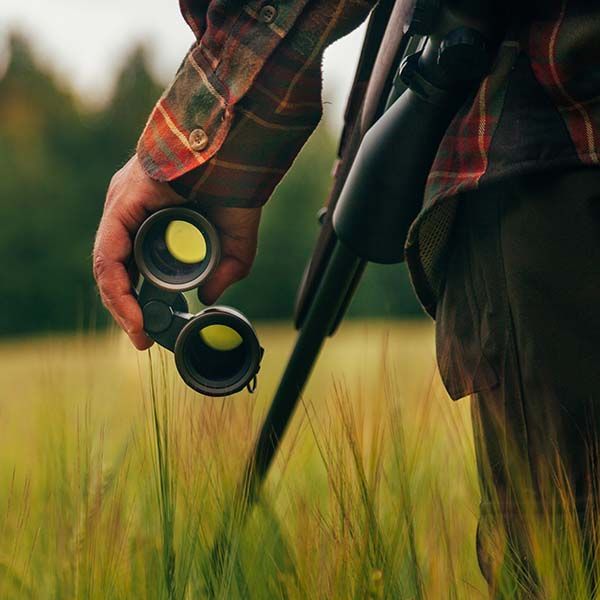 Person holding binoculars beside a tripod in a grassy field