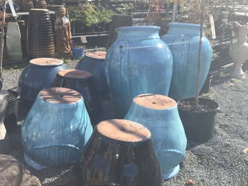 Blue ceramic jars and pots displayed outdoors on gravel, with plants and garden items in the background