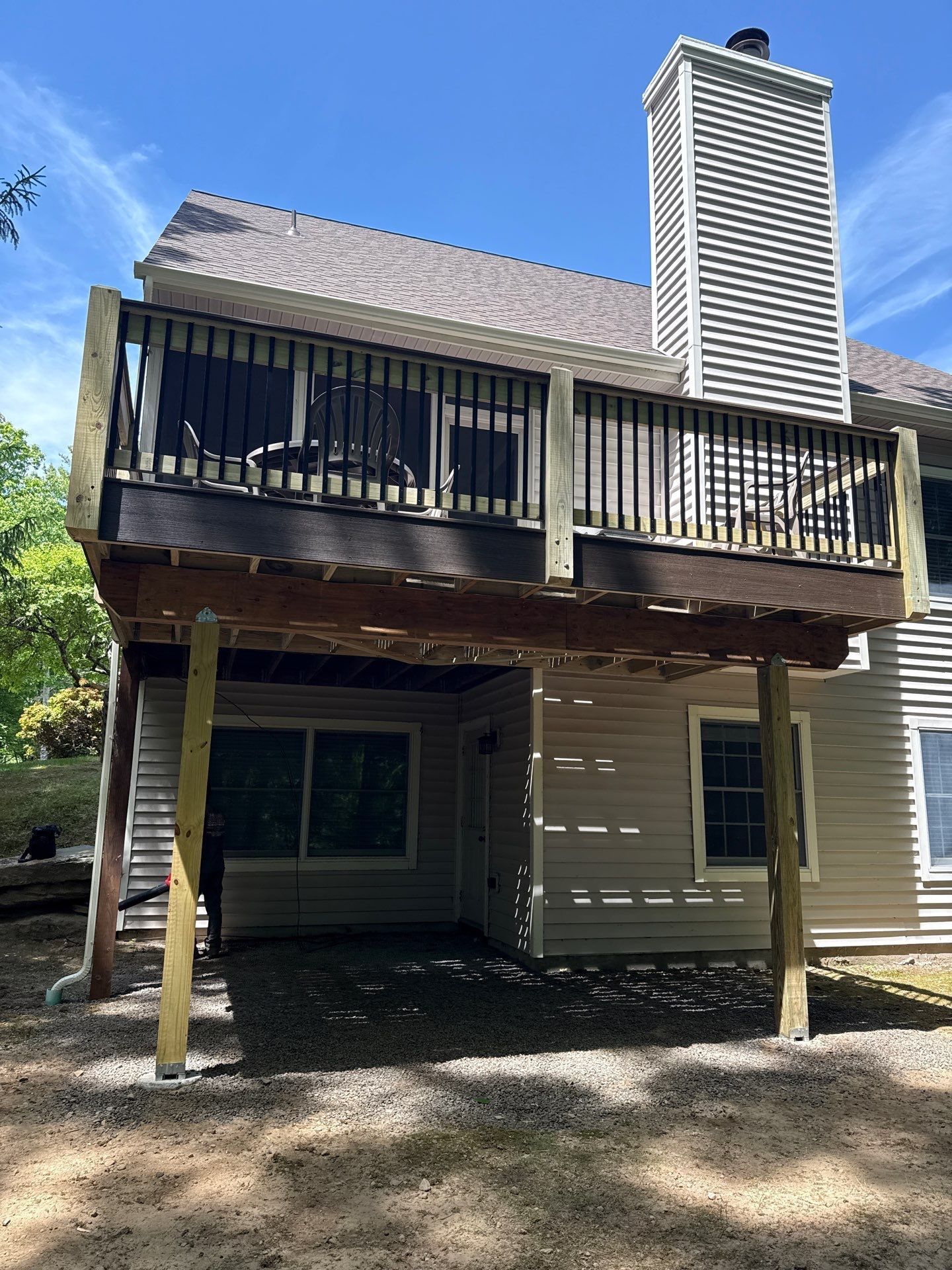 Back view of a beige house with a raised deck, wooden stairs, and a tall chimney in a wooded yard
