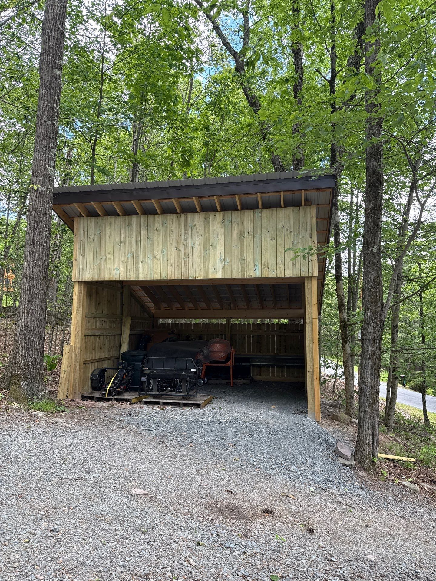 Wooden carport in the woods with a black vehicle parked inside and gravel driveway in front