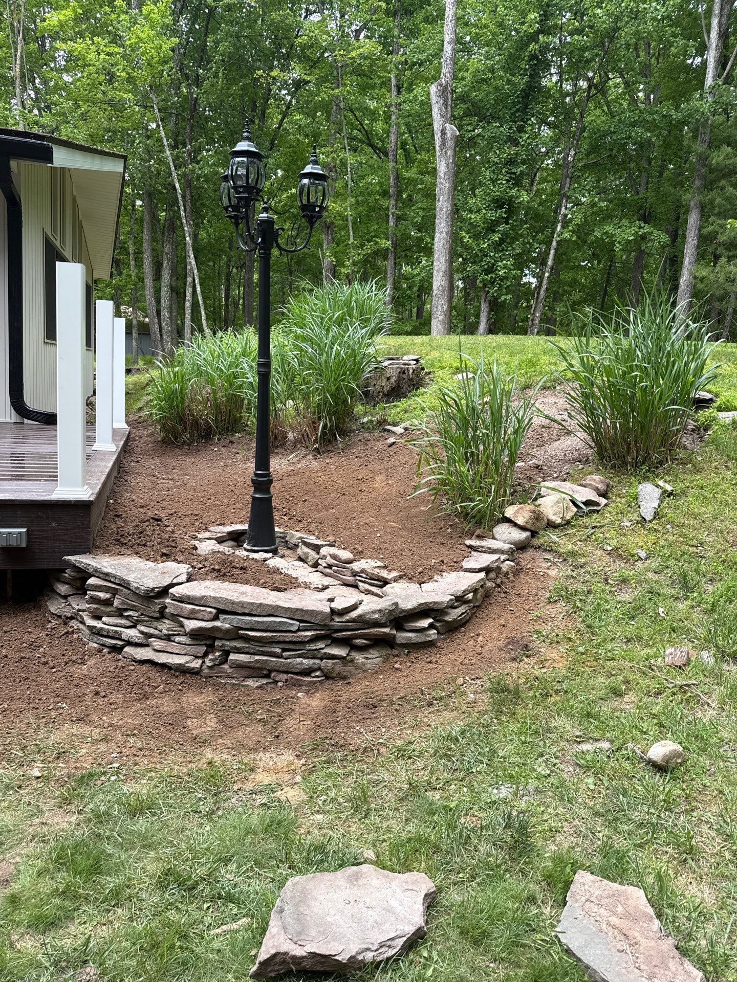 Backyard garden with stone border, black lamp post, shrubs, and a white porch at the edge of a wooded lawn