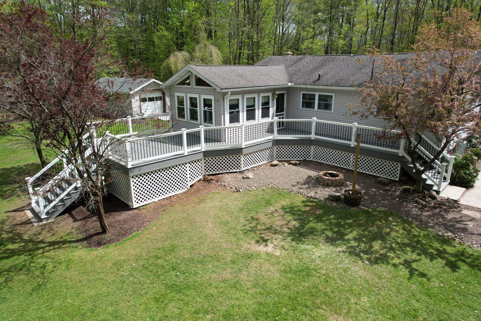 White house with a wraparound deck and stairs, surrounded by green lawn and trees