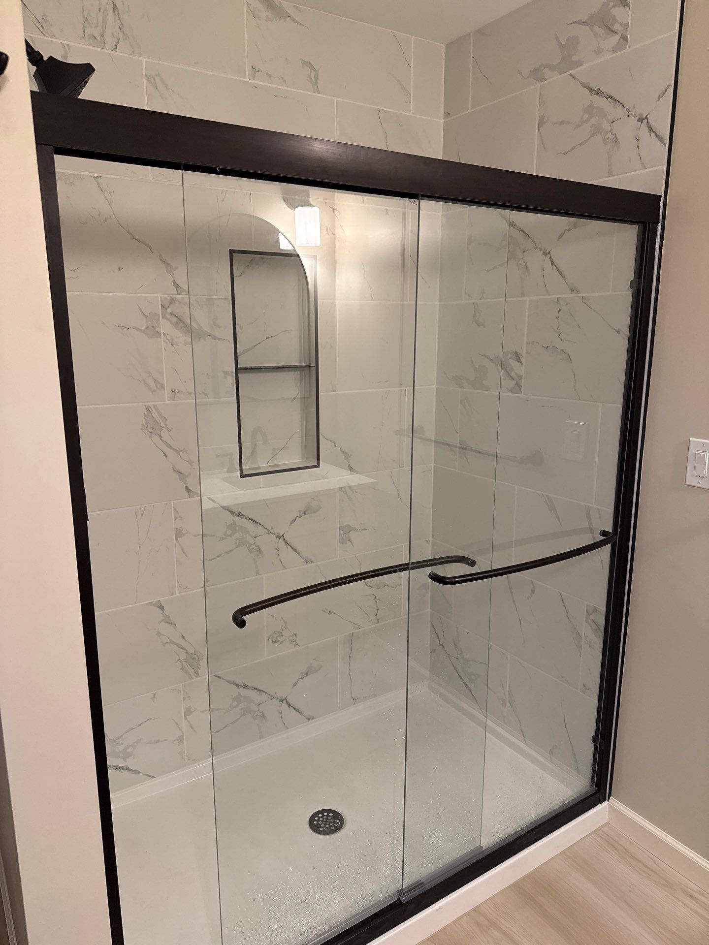 Glass shower enclosure with black frame and white marble tile walls, featuring a built-in niche and grab bar.
