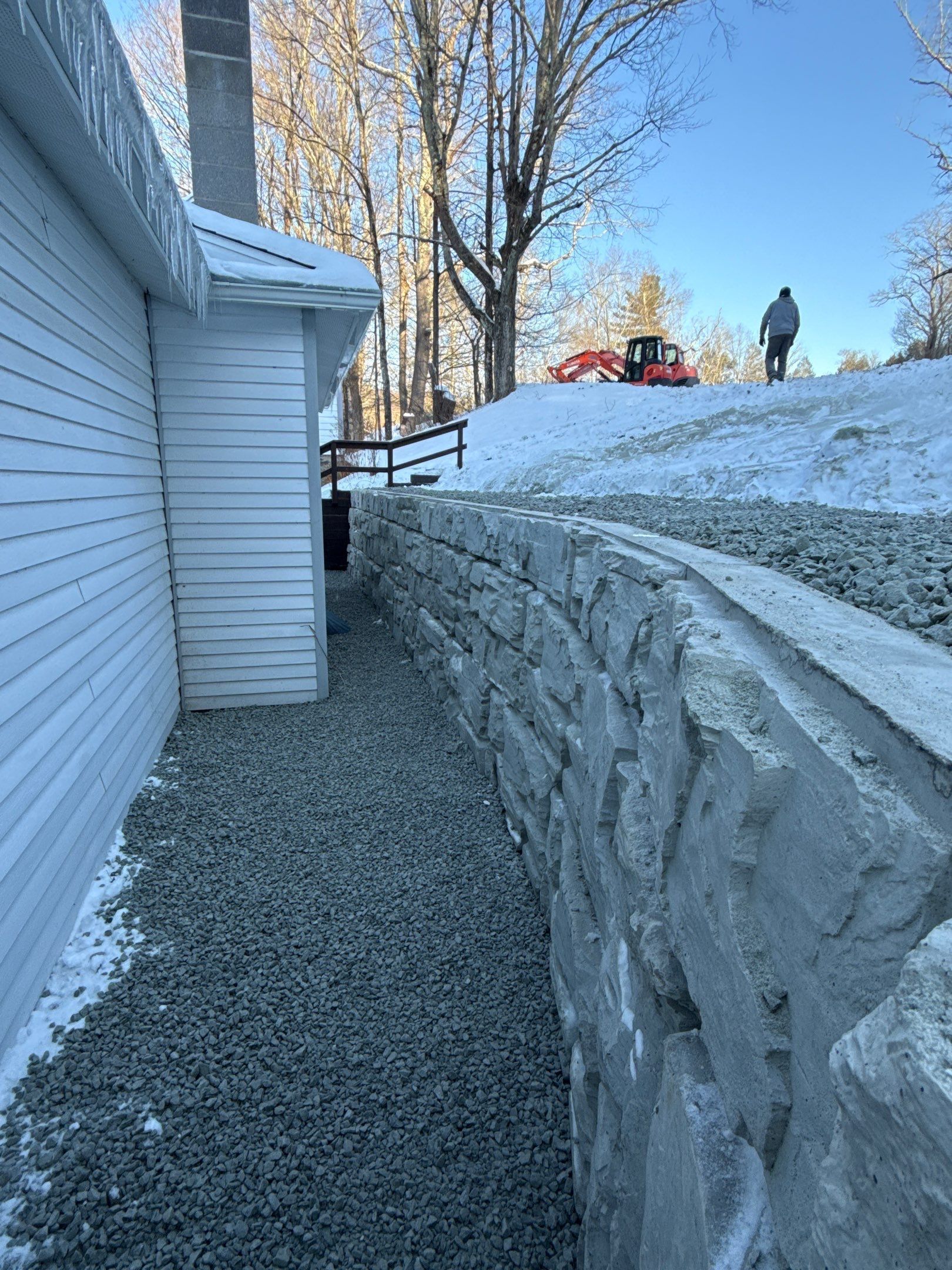 Narrow snowy side yard beside a white house, with a stone retaining wall and a person on the hill above.