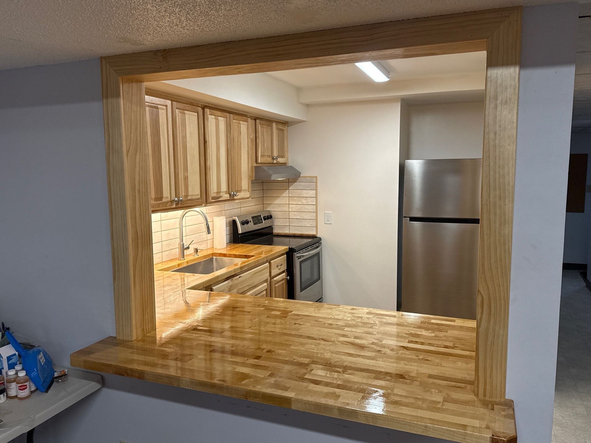 Small kitchen seen through a wide pass-through window, with wood cabinets and a stainless-steel refrigerator.
