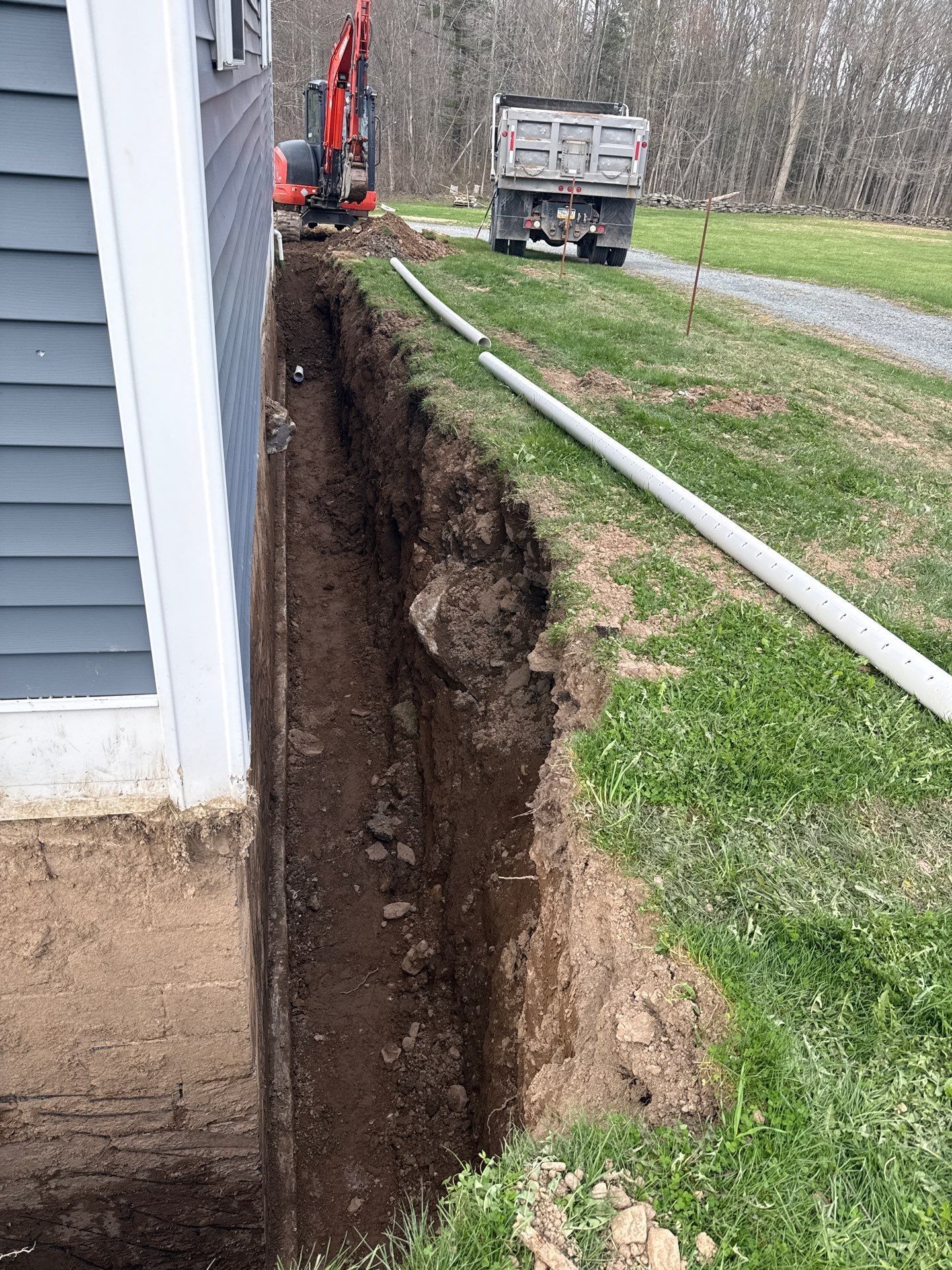 Deep trench dug beside a house, with an excavator and dump truck in the background.