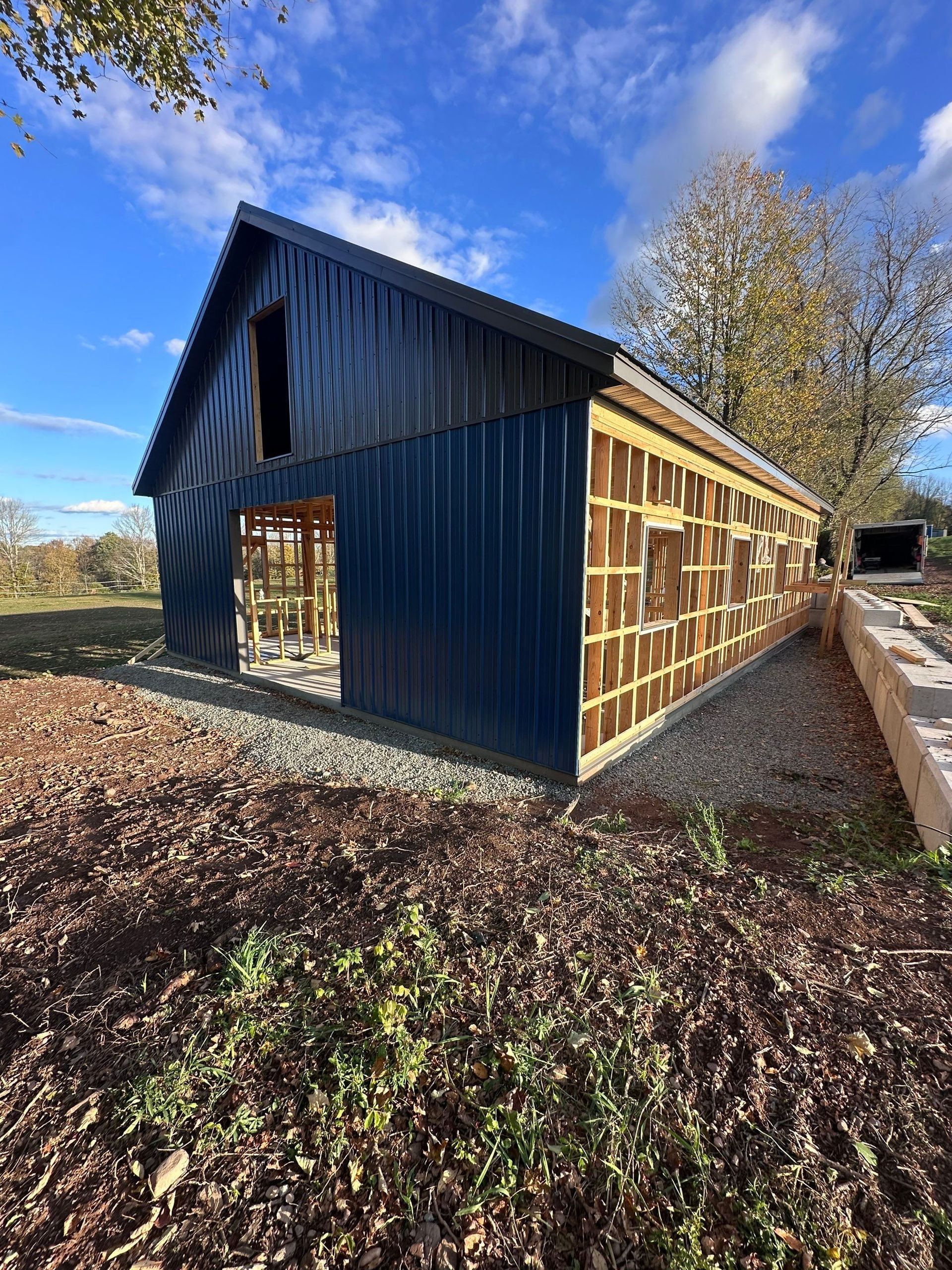Dark blue wooden barn with exposed framing on a rural dirt path under a sunny blue sky