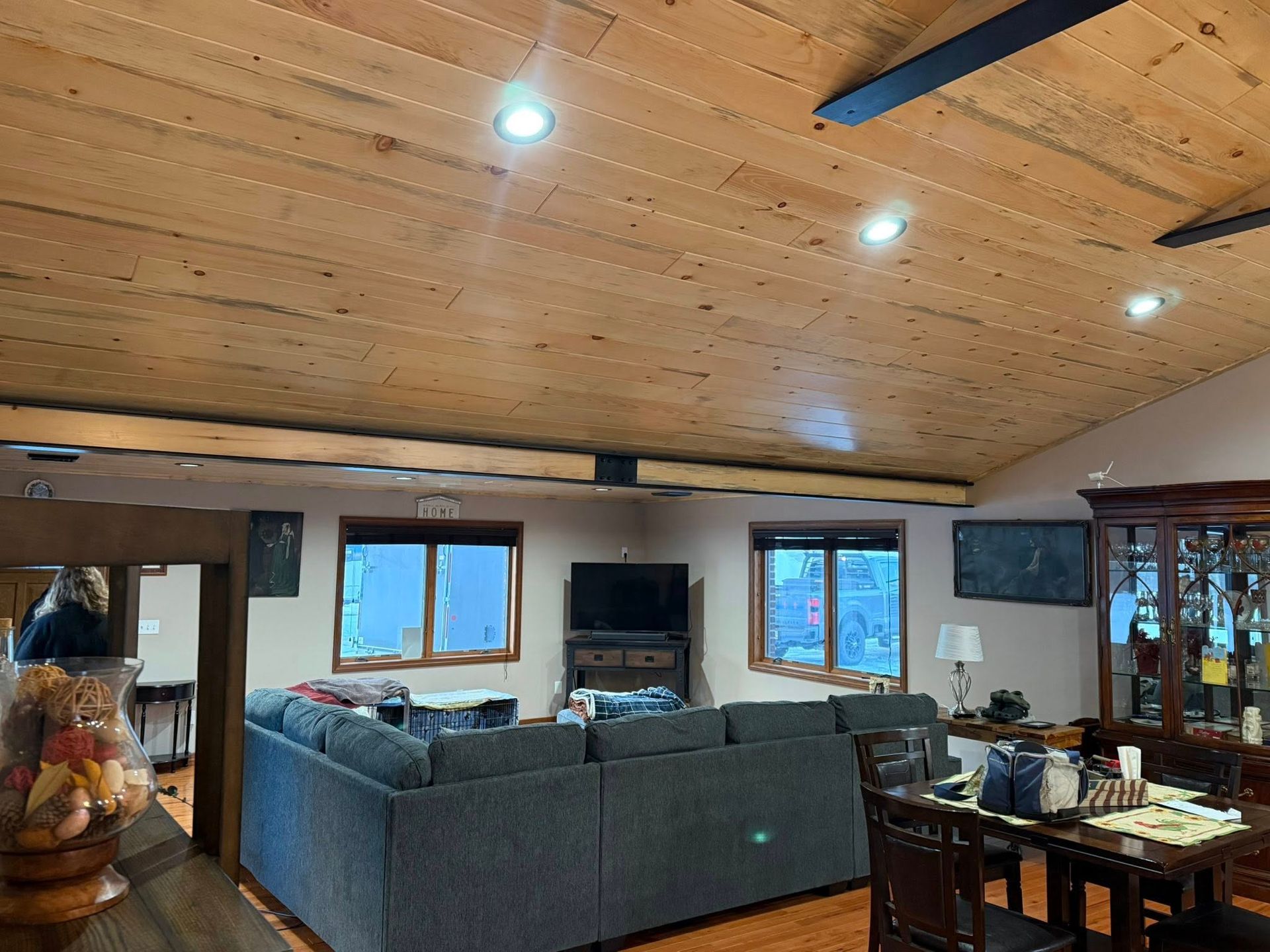 Cozy living room with wood ceiling, gray sectional sofa, large windows, and dining table on the right