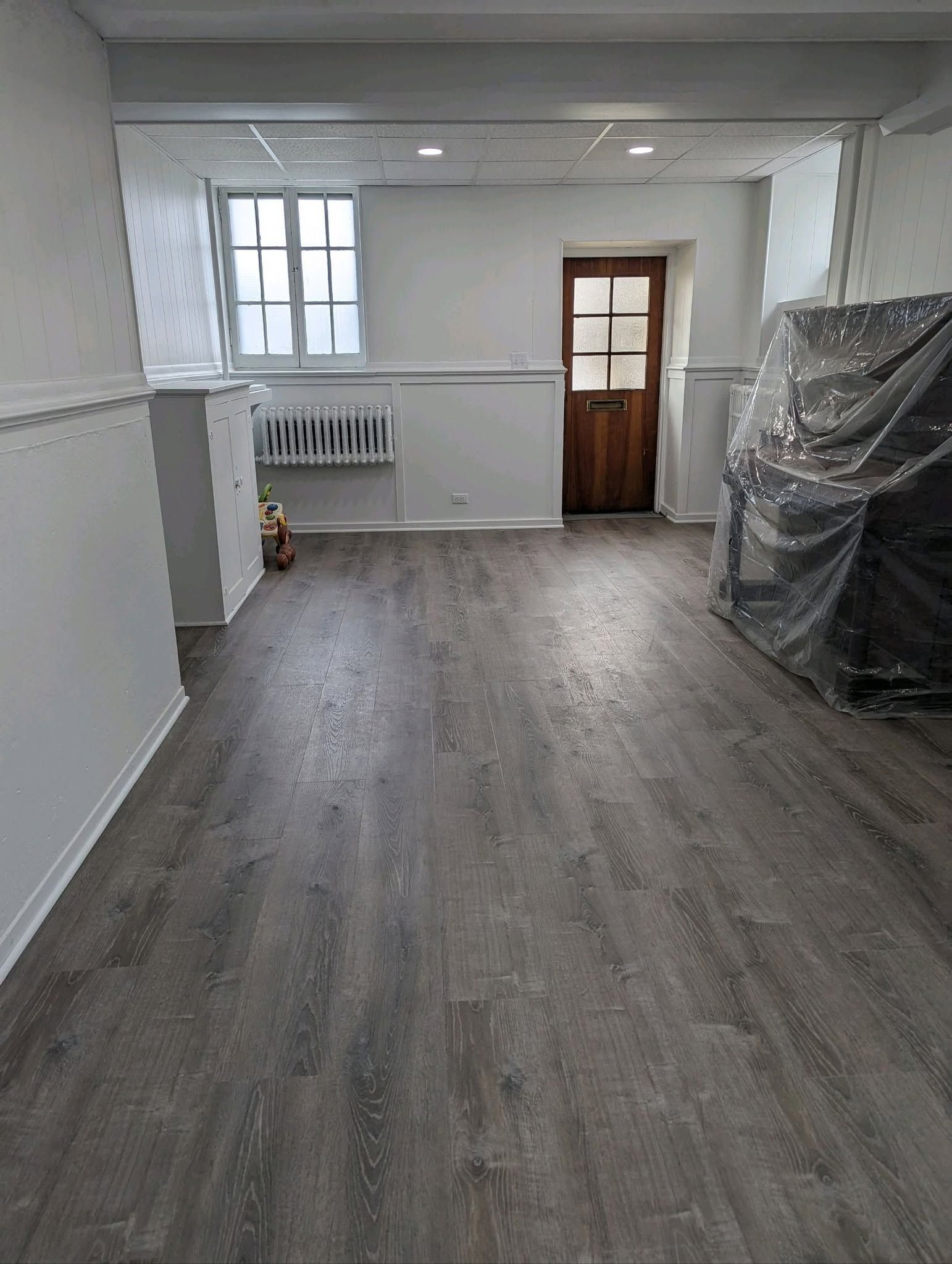 Empty room with gray wood flooring, white walls, a window, and a wooden door at the far end.