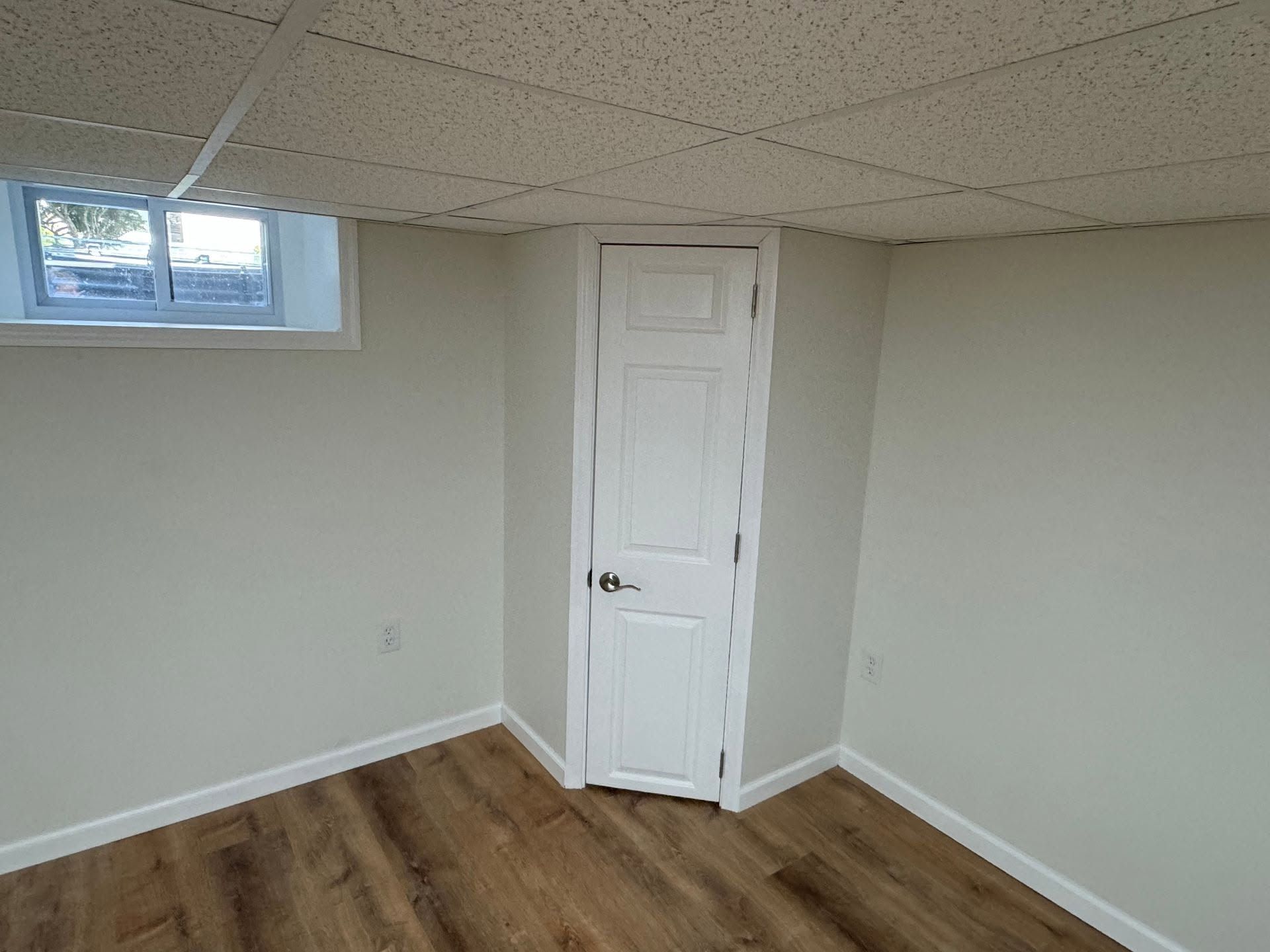 Empty room with wood floor, white walls, small window, and a white door in the corner