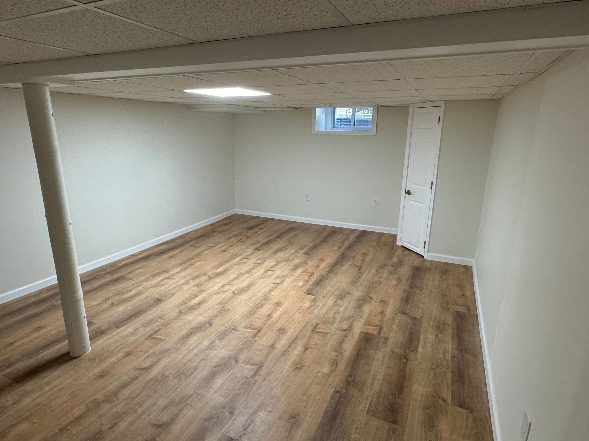 Empty basement room with wood flooring, white walls, a support column, and a small window.