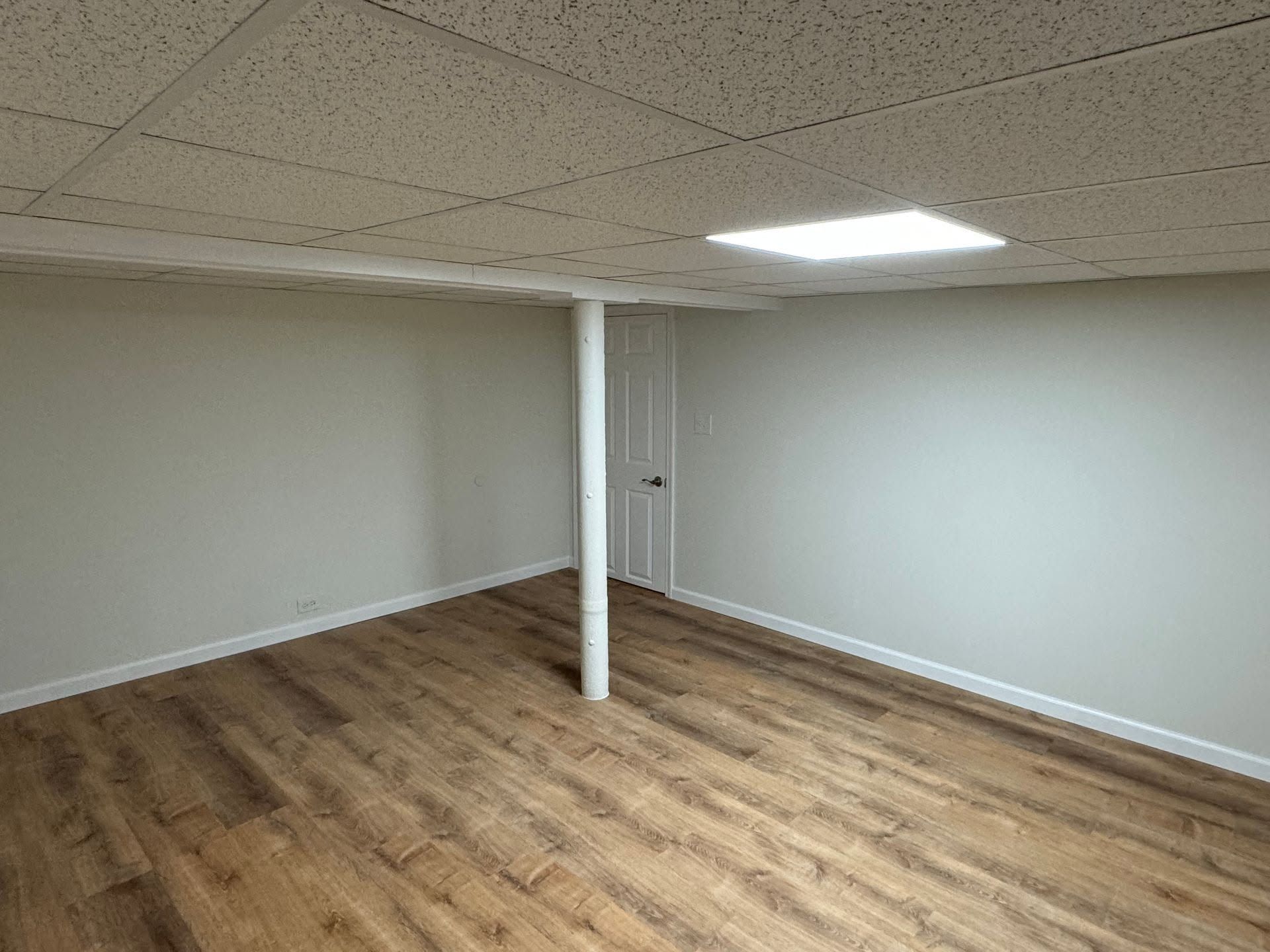 Empty basement room with wood-look floor, white walls, drop ceiling, and a support pole