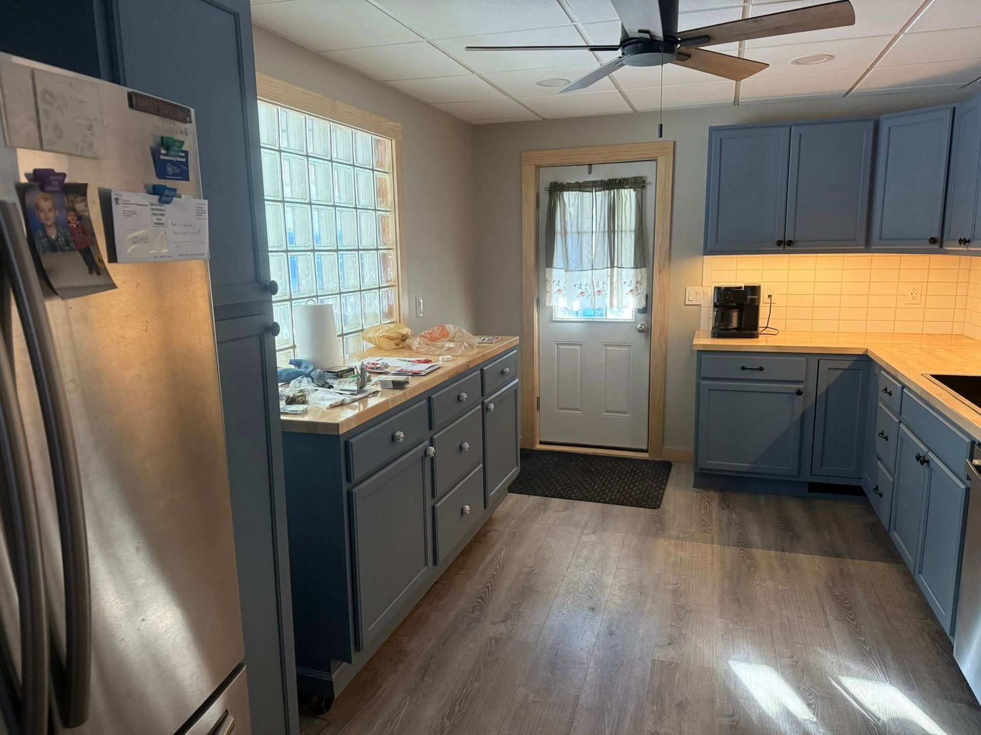 Bright kitchen with blue-gray cabinets, stainless fridge, wood floor, and window over the sink.