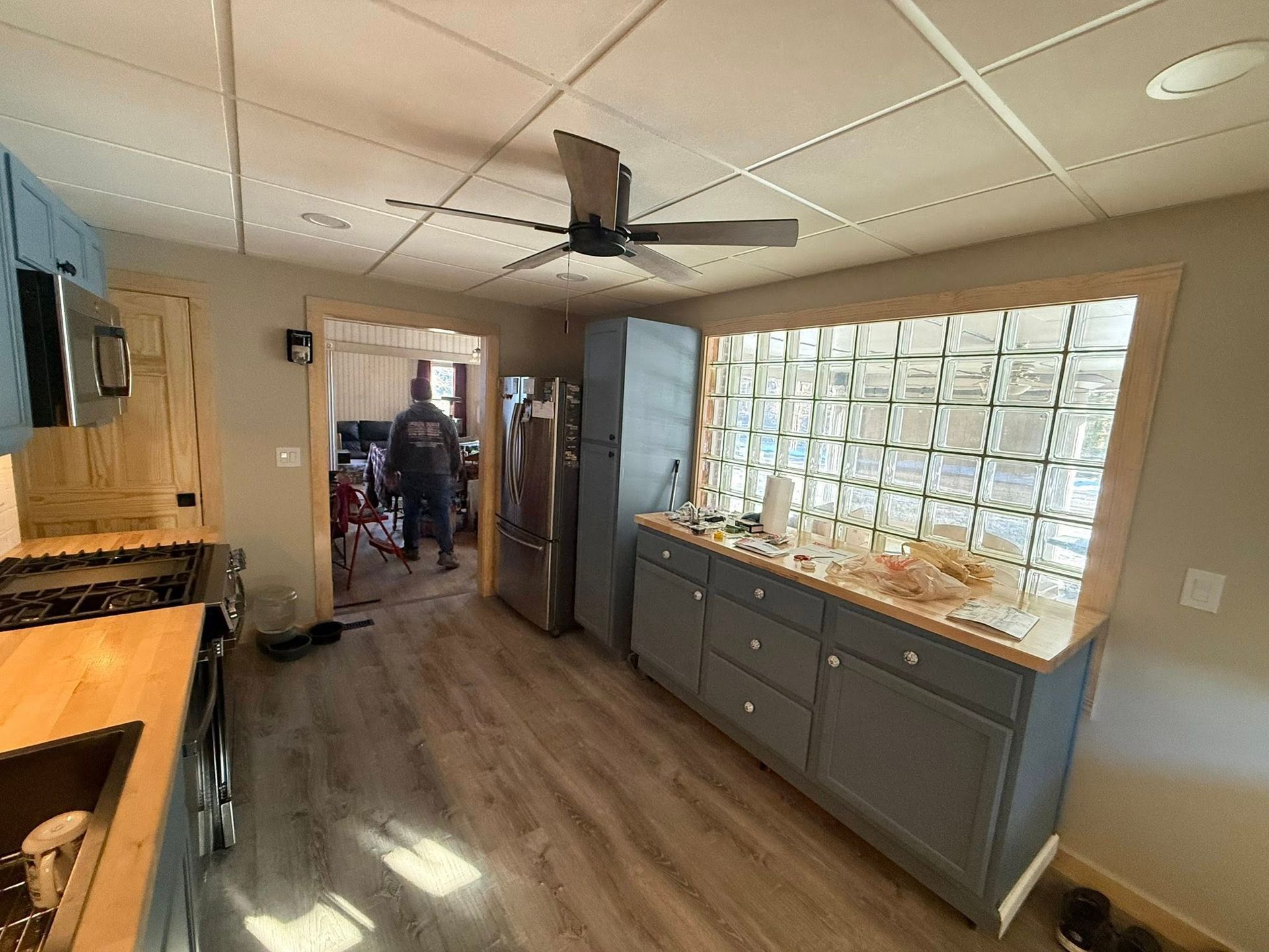 Bright kitchen with island, gray cabinets, glass-block wall, ceiling fan, and a person in the doorway.