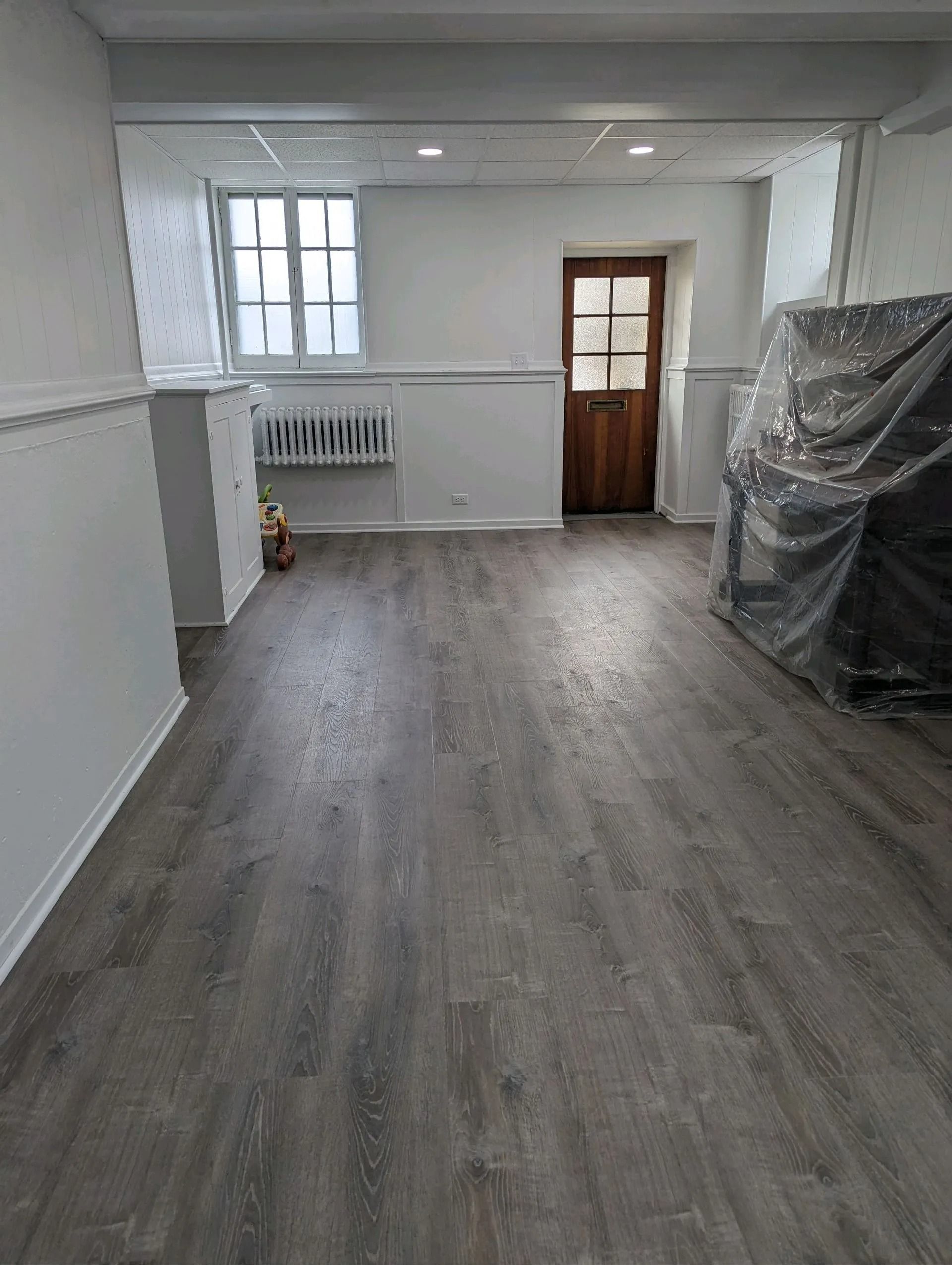 Empty room with gray wood floors, white walls, a window, and a wooden door at the back.