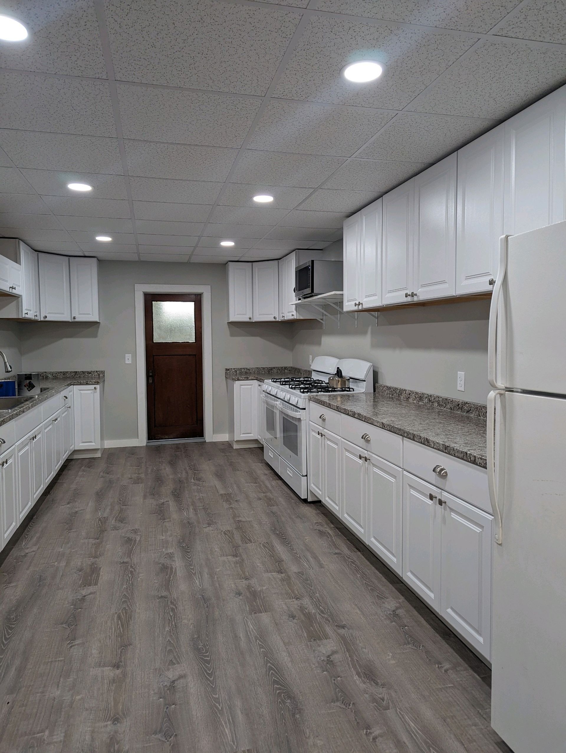 Long, empty white kitchen with white cabinets, gray flooring, and a wooden door at the far end