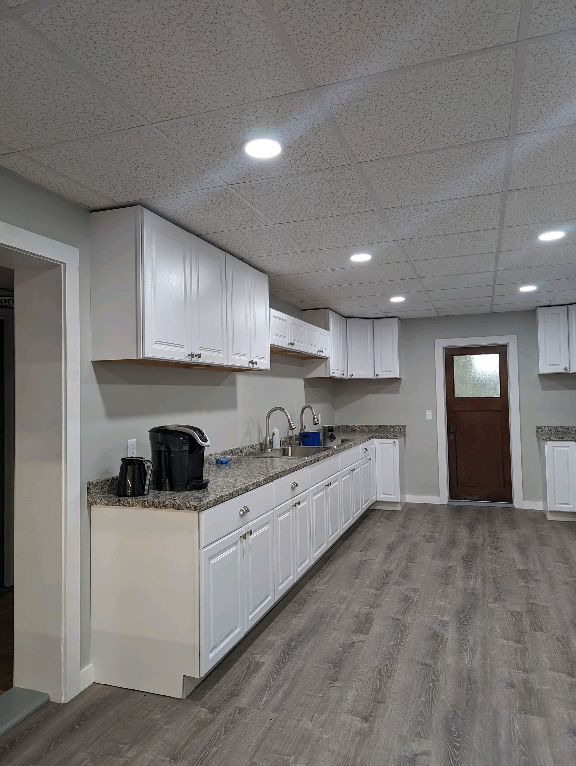 Bright white kitchen with cabinets, countertop appliances, and gray wood flooring leading to a door.