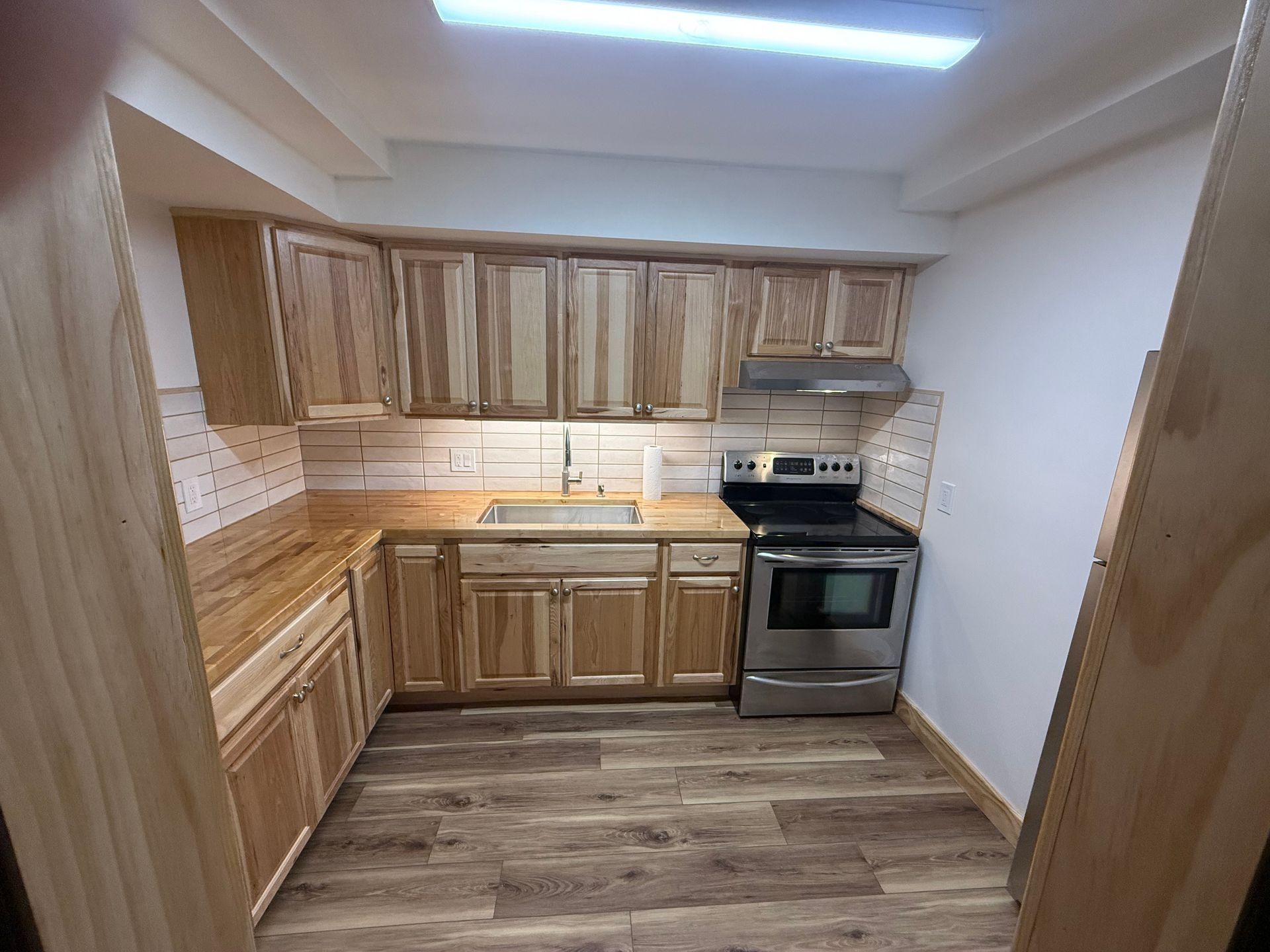 Small empty kitchen with light wood cabinets, stainless stove, and wood-look flooring.