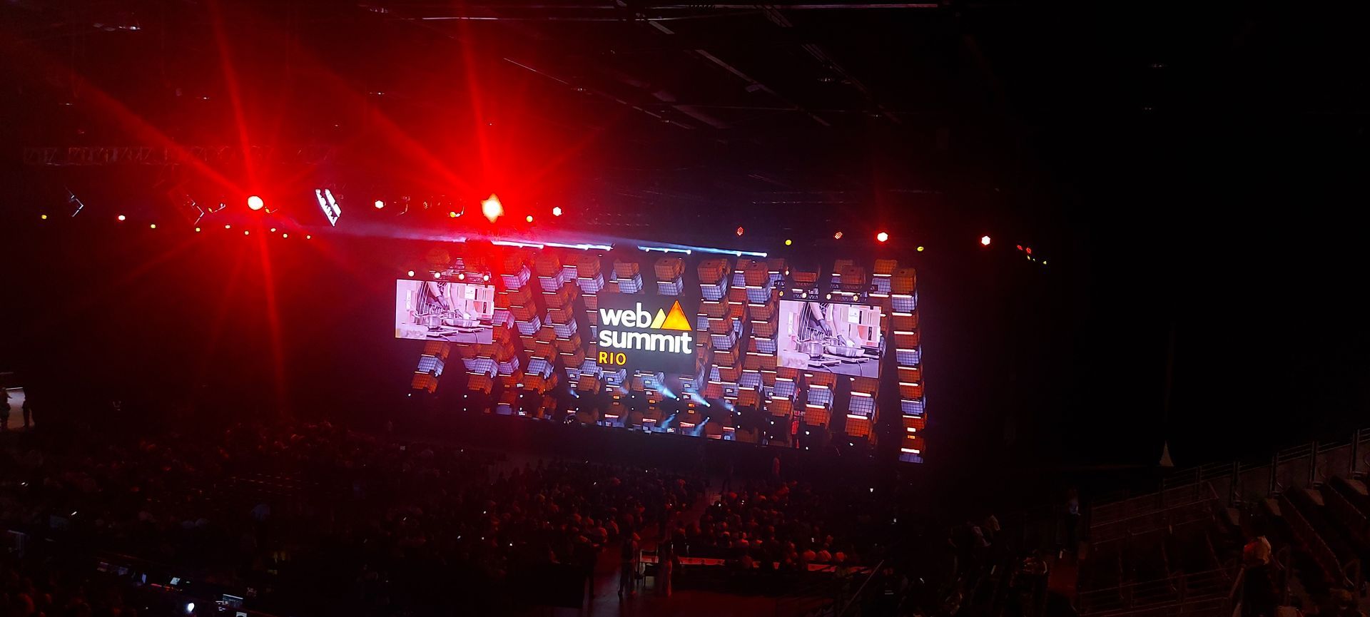 Red-lit concert stage with a large video screen and crowd in a dark arena