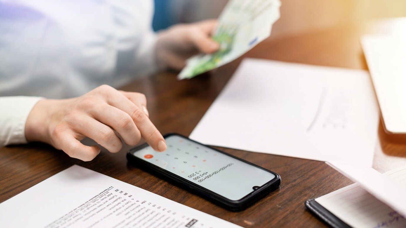 Hand using smartphone beside paperwork on a desk, with cash in the background
