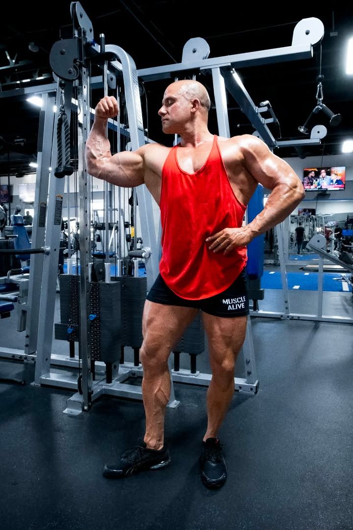 Muscular man in red tank top posing in a gym beside cable machine