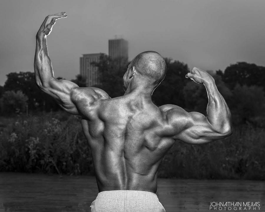 Black-and-white photo of a muscular back-flexing bodybuilder outdoors by water at dusk.